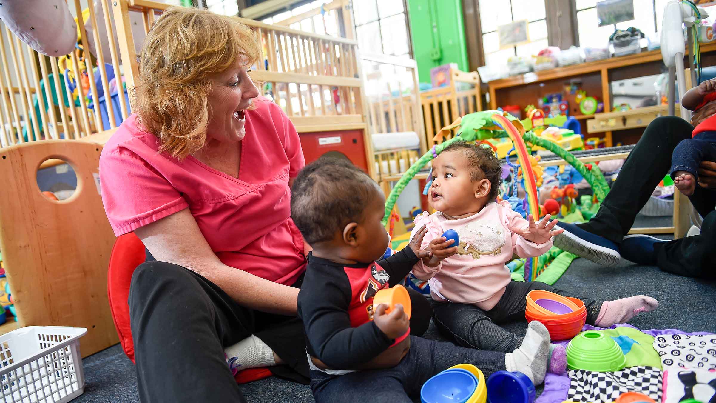 An early childcare teacher is smiling at two babies that are playing on the ground with toys