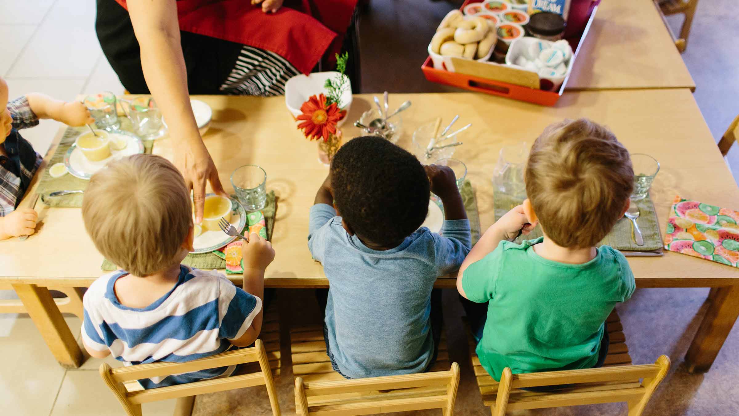 A group of preschoolers sit together at a table and are being served a snack